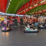 Kolorowe samochodziki na torze w wesołym miasteczku z barwnym tłem Formuły 1. Zielono-czerwony dach. Bawiące się dzieci i dorośli.