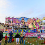 Kolorowy, wesoły lunapark z różowymi i zielonymi elementami. Na pierwszym planie karuzela, w tle tłum ludzi. Obok domy i drzewa.