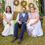 Trzy osoby siedzą na snopku siana, tło z kolorowymi kwiatami i wieńcami. Na ziemi zielona trawa, widoczny biały namiot. Dominuje kolor biały i kolorowe akcenty.