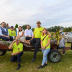 Grupa ludzi w jasnych koszulkach siedzi na kolorowym wozie zaprzężonym w traktor. Tło stanowią zielone pola i kilka samochodów. Obraz jest pełen jasnych, wesołych kolorów.