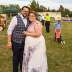 Para w eleganckich strojach na tle plenerowego wydarzenia, trawiaste podłoże. W tle namiot i ludzie. Kolory pastelowe, spokojne.