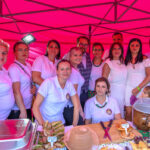 Grupa ludzi w białych ubraniach stoi pod różowym namiotem. Na stole przed nimi są różne potrawy. W tle jasne oświetlenie.