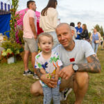 Na zdjęciu widoczny jest mężczyzna i małe dziecko na trawie. W tle ludzie i kolorowe namioty. Dziecko trzyma lizaka.