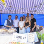 Grupa osób pod niebiesko-białym namiotem celebruje nagrody. Obok słodycze pod kloszami i kolorowe kwiaty. Tło w niebieskim odcieniu.