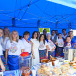 Grupa ludzi w strojach ludowych stoi pod niebieskim namiotem. Przed nimi stół z wypiekami i napojami. W tle widać zielone drzewa.