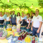 Grupa osób w kolorowych strojach stoi pod zielonym namiotem. Na stole znajdują się słoje z napojami i koszyk z lawendą. W tle girlandy i rośliny.