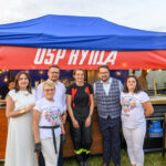 Grupa osób stoi przed niebiesko-czerwonym namiotem. Tło przedstawia stoisko z lampkami. Trawa pod stopami, dzień.