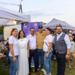 Grupa ludzi stoi na trawie, na tle namiotów w pasy. Mają na sobie casualowe ubrania w jasnych kolorach. Scena na zewnątrz, święto plenerowe.