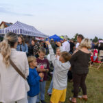 Grupa ludzi na festynie na tle stoiska z biało-niebieskim zadaszeniem. Trawiaste podłoże, w tle sceny z kolorowymi światłami. Dzień jest pogodny, niebo jasne.