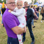 Mężczyzna w fioletowej koszulce trzyma dziecko na ramionach. Dziewczynka nosi różowe opaski na uszy. W tle plenerowe wydarzenie.