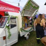 Biały pojazd z zielonymi dekoracjami stoi przy czerwonym namiocie. W tle ludzie w kolorowych ubraniach. Zielone trawiaste podłoże.