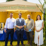 Czworo osób stoi przed żółtym namiotem z napisem „Miód z rodzinnej pasieki”. Tło jest zielone, z roślinami kukurydzy. Mężczyźni ubrani elegancko, kobieta w białej sukni.