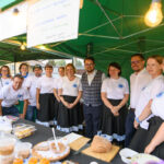 Grupa osób w białych koszulkach stoi pod zielonym namiotem. Na stole widoczne jedzenie, chleb. Tło zielone, oświetlone.