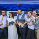 Pięć osób trzyma drewniane figurki na tle niebieskiego namiotu. Uśmiechnięci, ubrani elegancko, mają różnorodne rekwizyty. W tle widoczne kolorowe obrazy.