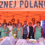 Grupa ludzi w strojach ludowych stoi pod pomarańczowym namiotem. Stoisko z jedzeniem i wieńcami z tyłu. Tło jest jednolite.