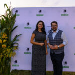 Dwóch ludzi stoi przed białą ścianką z herbem Gmina Dobre. Po lewej stronie rośliny. Tło jest jasne, trawiaste.