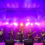 Zespół gra na scenie oświetlonej fioletowym światłem. Tło stanowi niebieski dach i czarna ściana. Na scenie są instrumenty i wokalista.