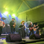 Zespół muzyczny występuje na scenie. Tło sceny jest zielone, oświetlone reflektorami. Muzycy grają na różnych instrumentach.