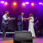 Scena oświetlona na różowo z trzema osobami rozmawiającymi na środku. Mężczyzna w garniturze podaje coś kobiecie w białej sukni. W tle widoczny napis na koszulce.