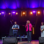 Uśmiechnięta grupka dzieci w kaskach strażackich, trzyma wąż strażacki za nimi stoją strażacy w mundurach.