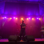 Zespół występuje na scenie oświetlonej fioletowym i pomarańczowym światłem. Na przodzie trzech muzyków: gitarzysta, wokalista, klawiszowiec. Tło to ciemny teatr pod namiotem.