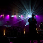 Na scenie koncertowej widać dwóch muzyków w cieniu, oświetlonych fioletowym i niebieskim światłem. W tle widoczne są reflektory. Atmosfera jest dynamiczna i intensywna.