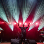 Zespół gra na scenie oświetlonej kolorowymi reflektorami. Dominują czerwone i niebieskie światła. W tle perkusja i syntezator.