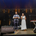 Na scenie pod niebieskim dachem stoi mężczyzna w kamizelce i kobieta w białej sukni z mikrofonem. Tło jest ciemne z pomarańczowymi światłami i instrumentami. Obok stoi czerwony keyboard na statywie.