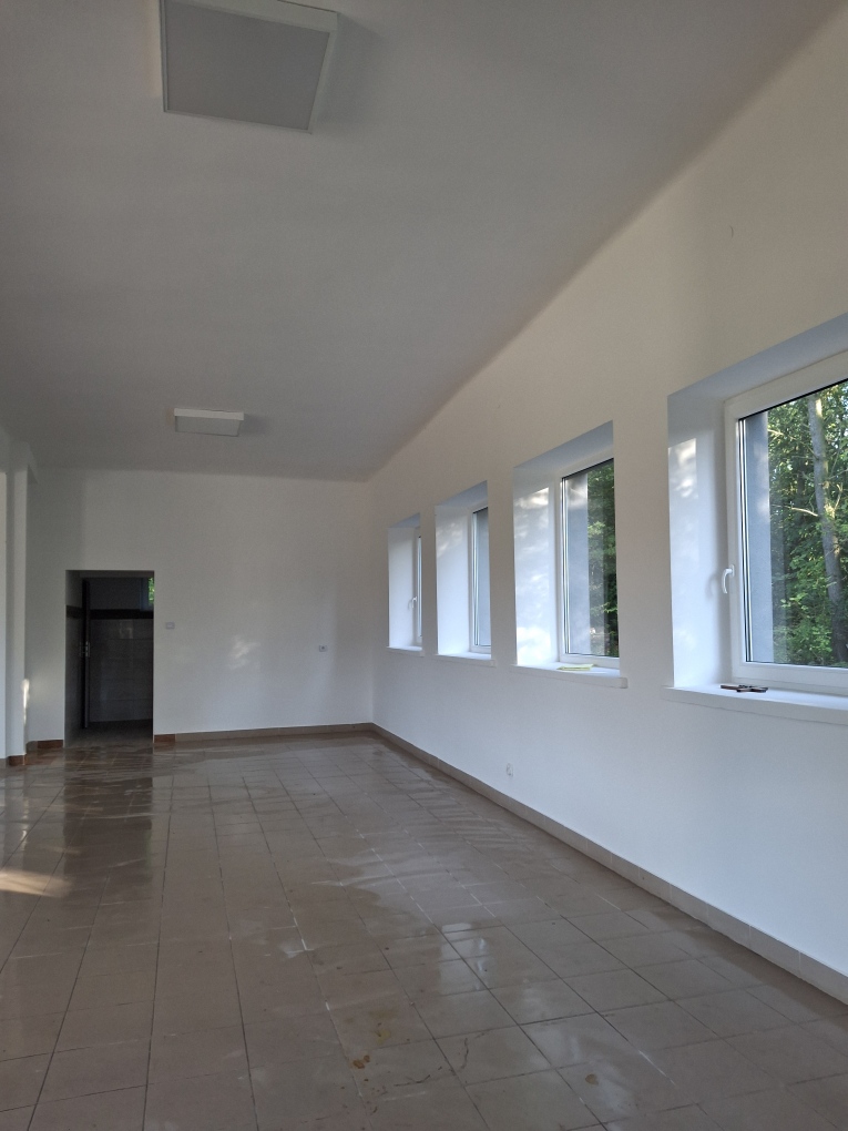 Puste pomieszczenie z białymi ścianami i dużymi oknami. Podłoga pokryta jasnymi płytkami, przez które przepuszczane jest światło. Na zewnątrz okien widać drzewa.