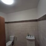 Jasna łazienka z białą toaletą i umywalką, ściany wykończone beżowymi płytkami. Okrągłe źródło światła na suficie. Drzwi drewniane po lewej stronie.