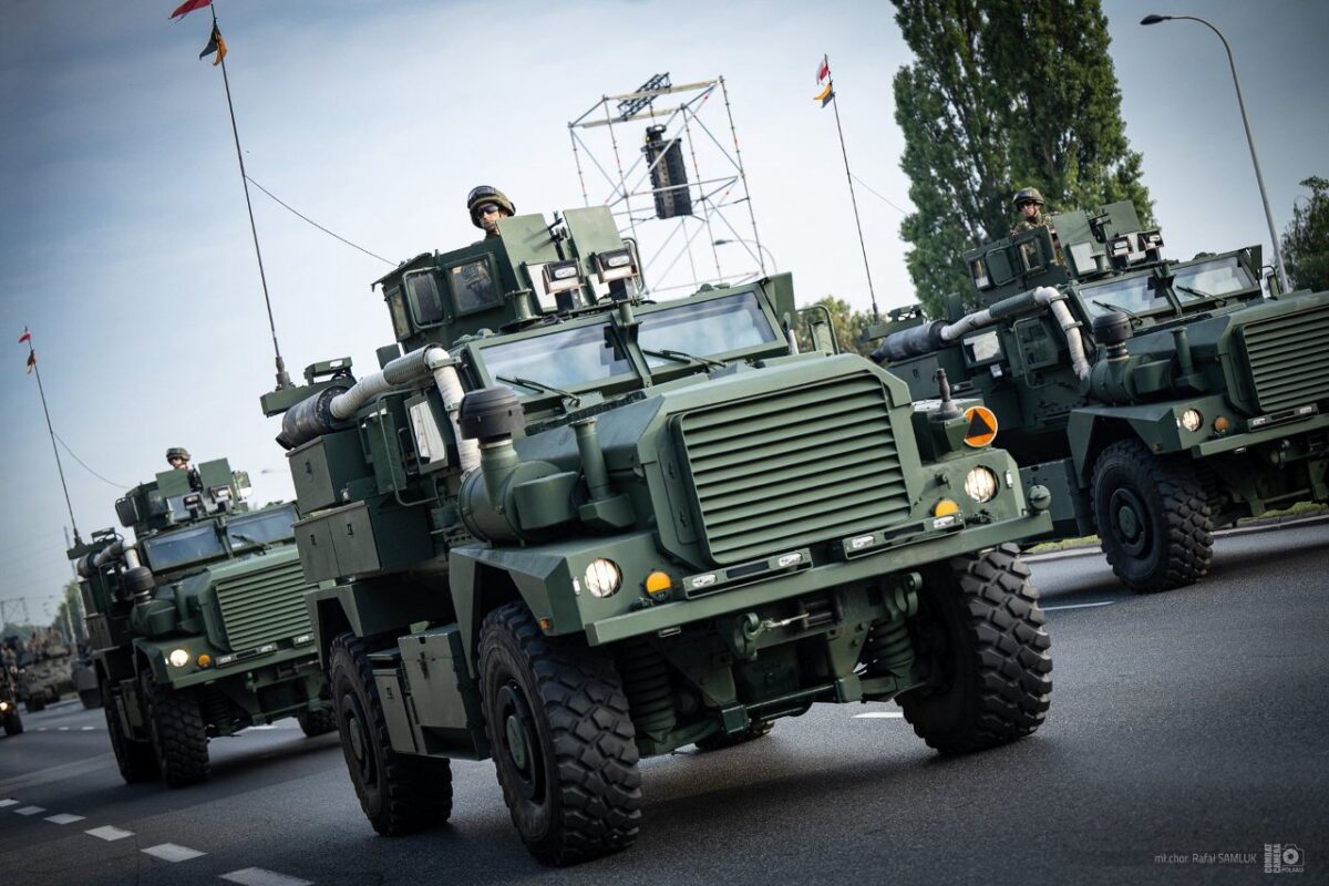 Zielone pojazdy wojskowe jadą po drodze. W tle widoczne drzewa i maszt z flagami. Niebieskie niebo i żołnierze w kaskach.