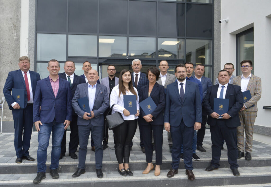 Grupa ludzi w formalnym ubraniu stoi na schodach przed dużym oknem. Trzymają niebieskie teczki. Tło to szklana fasada budynku.