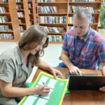 Dwoje ludzi pracuje przy stole w bibliotece. Kobieta pisze na zielono-żółtym formularzu. W tle widoczne są półki z książkami.