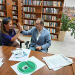 Na zdjęciu dwoje ludzi przybija piątkę przy drewnianym stole w bibliotece. Tło to regały z książkami i zielone rośliny. Na stole leżą ulotki i dokumenty, w tym oznaczony tekst Uniwersytet Dobrego Wieku