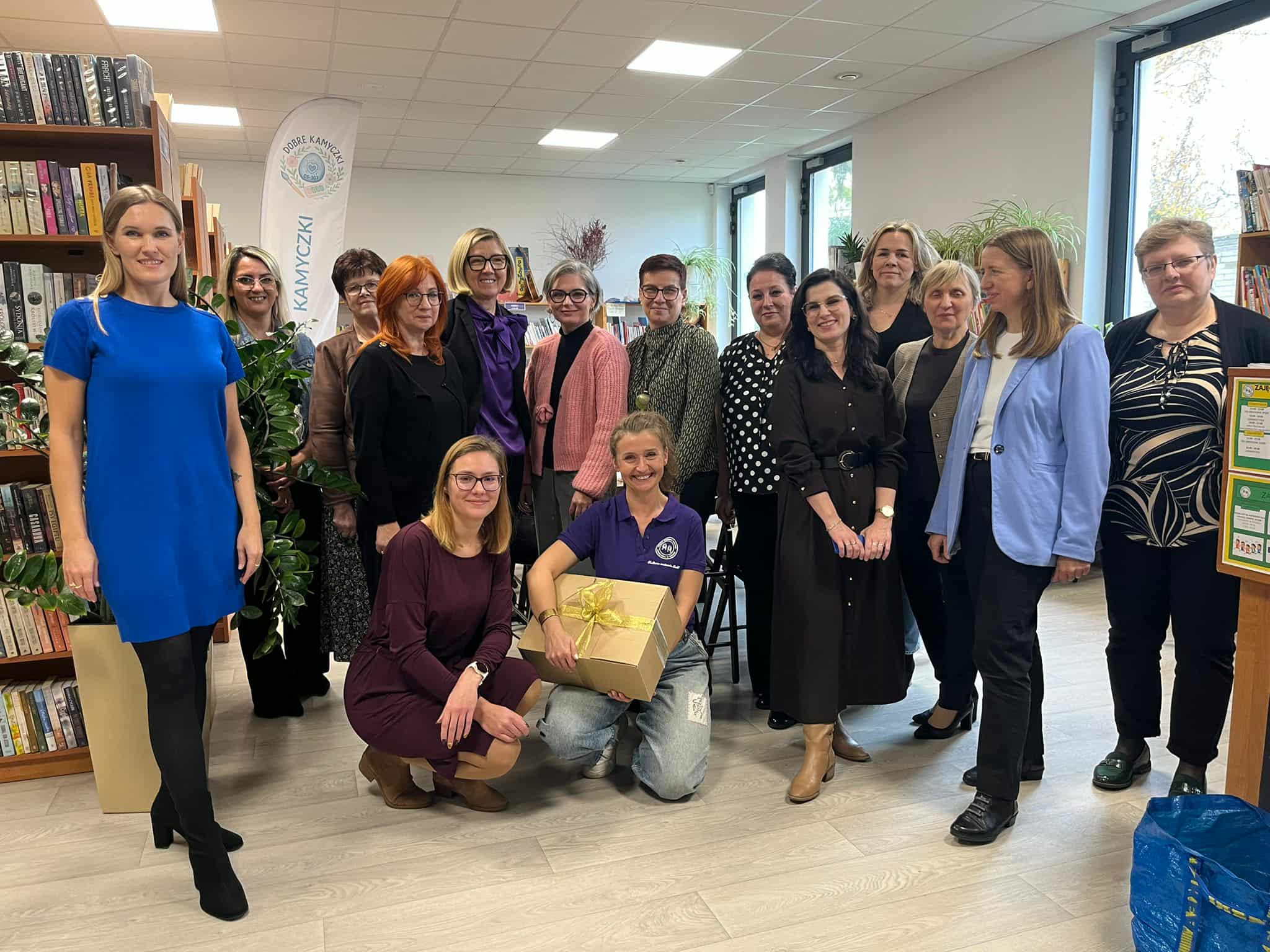 Grupa osób uśmiechniętych w bibliotece, stoją przed regałem z książkami. Osoba w niebieskiej sukience po lewej, reszta w eleganckich strojach. Jedna osoba klęczy z pudełkiem przewiązanym wstążką.