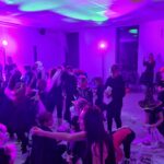 Widoczne jest halloweenowe przyjęcie dla dzieci w kolorowych kostiumach. Sala oświetlona na fioletowo z dekoracjami i papierem na podłodze. W tle są rodzice robiący zdjęcia.