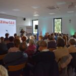 10 Sala z widownią siedzącą na krzesłach, w tle biały ekran z napisem Mazowsze. Osoby skupione na prezenterce obok mikrofonów.