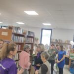 Grupa rozmów w bibliotece z półkami pełnymi książek. Jasne oświetlenie, tło z oknami i plakatami. Ludzie w kolorowych ubraniach.