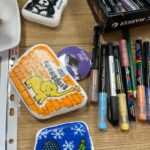 Na stole leżą pomalowane kamienie z motywami psa, słonia i zimowego krajobrazu. Kolory to czarny, pomarańczowy i niebieski. Obok znajduje się pudełko z pisakami.