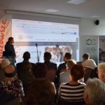 11 Osoba przemawia na tle prezentacji z logiem Uniwersytetu. Na scenie mikrofony, wokół siedzi publiczność. Z boku banery i światło z okna.