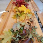 Na drewnianym stole leżą różnokolorowe jesienne liście: żółte, czerwone i zielone. Tło stanowią książki na półkach i szare fotele. Pomieszczenie jest jasne i przestronne.