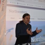 12 Osoba przemawiająca z mikrofonem na tle ekranu z napisem Northwestern. Po lewej biało-czerwona flaga.