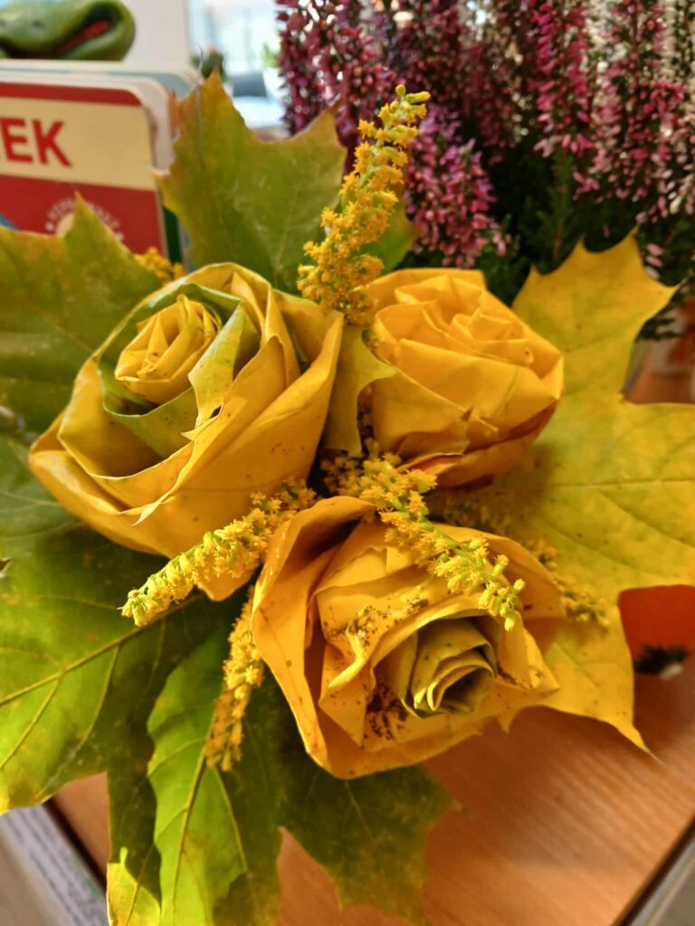 Bukiet z żółtych liści uformowanych w róże, przyozdobiony żółtymi kwiatami. W tle fioletowe wrzosy. Całość leży na drewnianym stole.