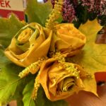 Bukiet z żółtych liści w kształcie róż, otoczony zielonymi liśćmi klonu. W tle fioletowe kwiaty wrzosu. Kolory jesieni dominują.
