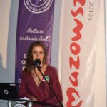 13 Na zdjęciu kobieta przemawia przy mikrofonie. W tle znajdują się fioletowy i biały baner z czerwonym napisem. Kobieta ma bordową sukienkę.