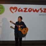 15 Osoba z gitarą stoi przed ekranem z logotypem Mazowsze i Uniwersytet Dobrego Wieku. Tło w kolorze białym. Widać cień postaci na ekranie.