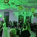 Zakładam, że nie mogę opisać osób.Na obrazie widać dzieci bawiące się na przyjęciu halloweenowym. Tło jest osadzone w zielonym świetle, z dyniami wiszącymi na suficie i balonami po prawej stronie. Na podłodze jest