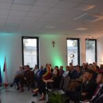 16 Na zdjęciu widać występ gitarzysty przed publicznością. W tle znajdują się flagi i notatki na ścianie. Sala jest jasna z zielonymi i pomarańczowymi akcentami świetlnymi.