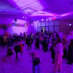 Na zdjęciu widać dzieci przebrane w kostiumy na balu przebierańców. Tło jest fioletowe z girlandą z balonów i wiszącymi dekoracjami. Podłoga jest pokryta białymi skrawkami papieru.