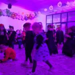 Dzieci w strojach Halloween bawią się w fioletowym oświetleniu. W tle balony i papierowe dynie na suficie. Na podłodze leżą rozrzucone papierki.
